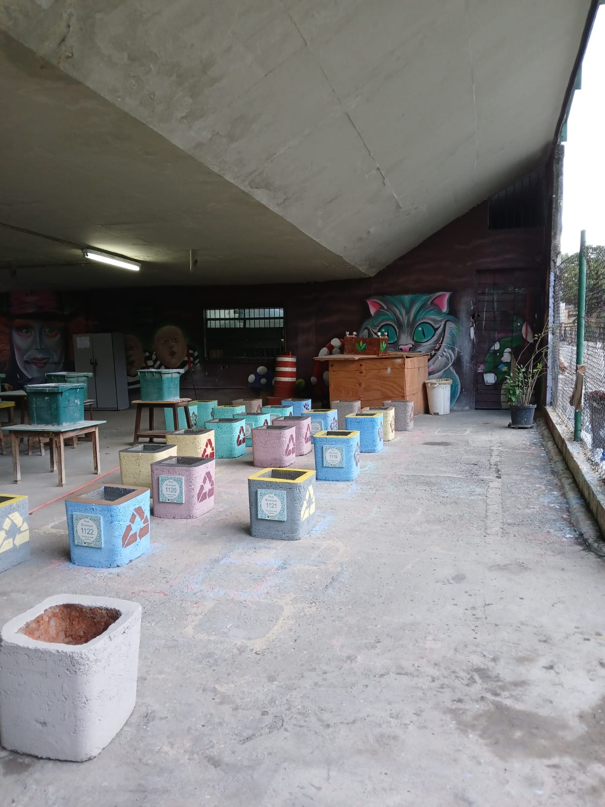 A imagem mostra um espaço coberto, possivelmente um galpão ou área sob um viaduto, com diversos blocos de concreto pintados em cores vibrantes, como azul, amarelo, cinza e rosa. Esses blocos possuem símbolos de reciclagem e números identificadores, sugerindo um projeto relacionado à sustentabilidade e descarte seletivo. Ao fundo, há uma parede com murais artísticos, incluindo um grande desenho do Gato de Cheshire, além de móveis e objetos que remetem a um espaço de trabalho ou oficina. O ambiente transmite uma atmosfera criativa e sustentável.